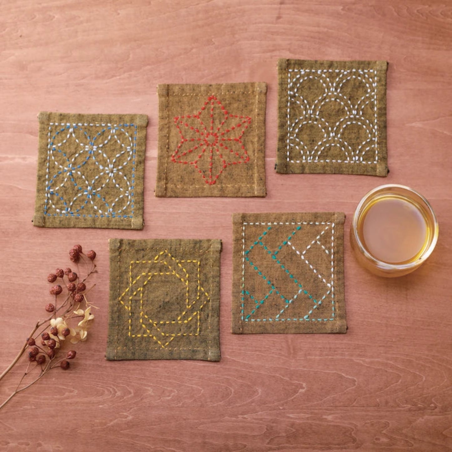 Five square linen coasters with colourful geometric and floral embroidery, arranged on a wooden surface next to a glass of tea and dried flowers.