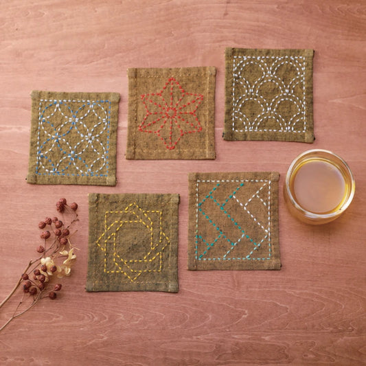 Five square linen coasters with colourful geometric and floral embroidery, arranged on a wooden surface next to a glass of tea and dried flowers.